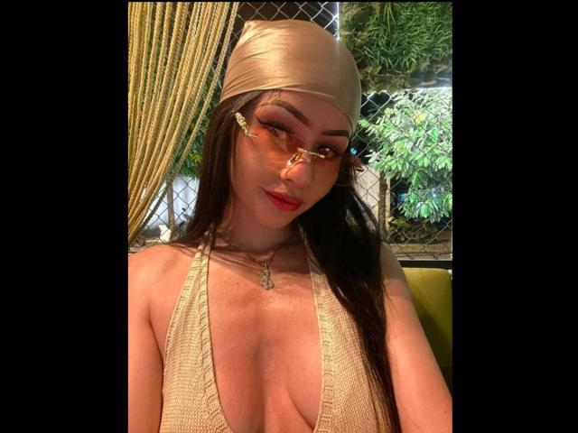 KattRouse with long brown hair, gold headband, and orange glasses, wearing a low-cut, beige top, showing her tits, with a green leafy background. Her red lips are slightly parted.