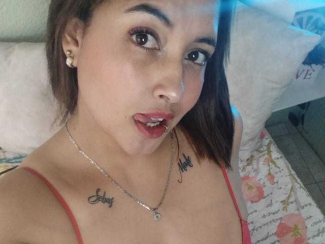 MirandaDreams with brown hair, red spaghetti strap top, earrings, necklace, tattoos saying "Love" and "Sky", parted lips, looking at camera, bedroom setting.