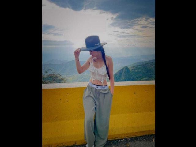 KattRouse stands in front of a yellow wall, wearing a white lace top, gray pants, and a black hat, with her hand on her head, smiling.