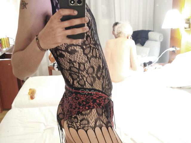 Teronn porte de la lingerie en dentelle noire avec un ruban rouge, montrant ses fesses nues et ses cuisses dans une photo prise devant un miroir. Une femme à la peau claire et aux cheveux courts, avec des cheveux gris, est nue en arrière-plan..