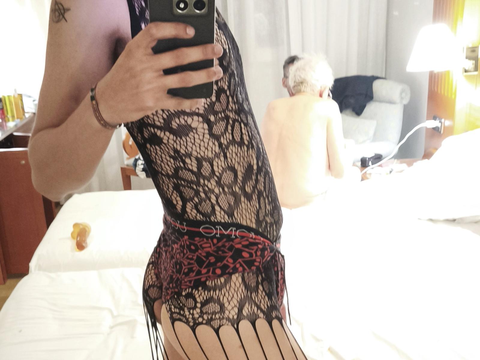 Teronn trägt schwarze Spitzendessous mit roter Schleife und zeigt ihr nacktes Gesäß und ihre Oberschenkel in einem Spiegel-Selfie. Eine kurzhaarige Frau mit grauem Haar ist im Hintergrund oben ohne..