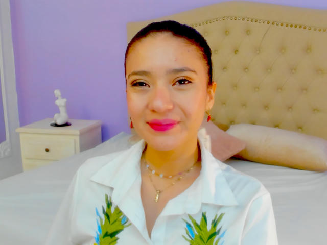 CattsyCathy with brown skin, black hair in a bun, pink lipstick, white shirt with green pineapple prints, necklace, smiling at camera. Bedroom with beige tufted headboard.