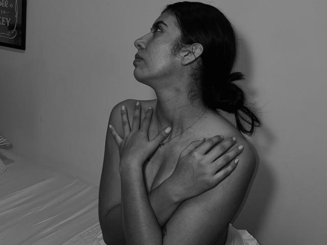 Black-and-white photo of MiluSky with hands on chest, topless, looking up, dark hair in a ponytail, small tits, fair skin, subtle freckles, nail polish visible, bed in background.