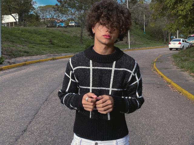 SteinMusic with curly hair, wearing a black sweater with white grid pattern, holding hands in front, standing on a curved road with a white car in the background.