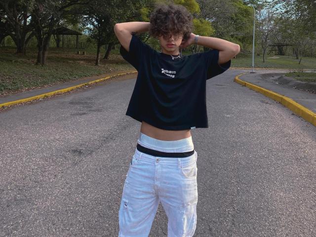 SteinMusic with curly hair, black crop top, and white pants standing on a road, hands behind head, exposing a black waistband. Road has yellow curbs, trees in background.