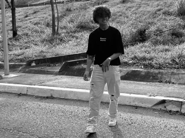 Black-and-white photo of SteinMusic with curly hair, wearing a dark shirt, light pants, and white sneakers, standing on a sidewalk, looking down, with a wristwatch on.
