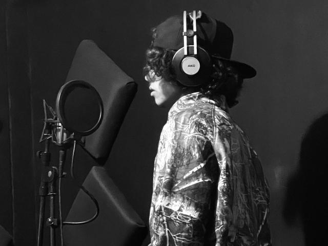 SteinMusic with curly hair, wearing a shiny, patterned jacket and headphones, stands in profile, facing a microphone on a stand, in a black-and-white shot.