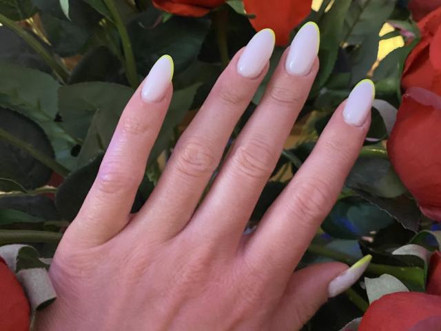 AlicePrings' hand with long, white, almond-shaped nails and neon yellow tips, surrounded by red roses and green leaves.