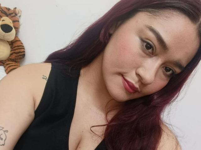 MaddieMoon con cabello morado, camiseta sin mangas negra, lápiz labial rojo, tatuaje en el brazo superior, sonrisa suave, ojos marrones, pechos pequeños, piel clara, un tigre de peluche a la izquierda..