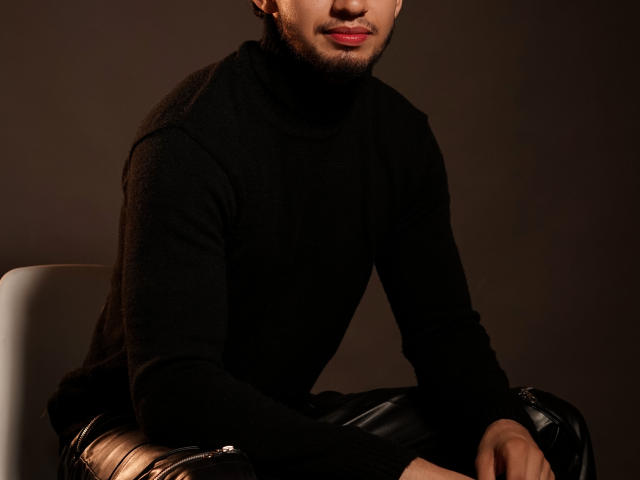 MichaelMagno with a black turtleneck, leather pants, and a beard, sitting on a chair, showing a smooth, slightly visible ass, and a cute, sexy, subtle smile.