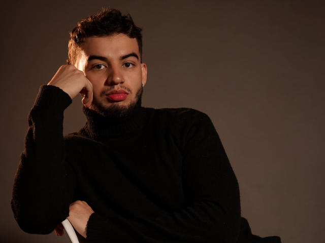 MichaelMagno, a cute dude with curly dark hair and a beard, wearing a black turtleneck, rests his chin on his hand, looking at the camera with red lips.
