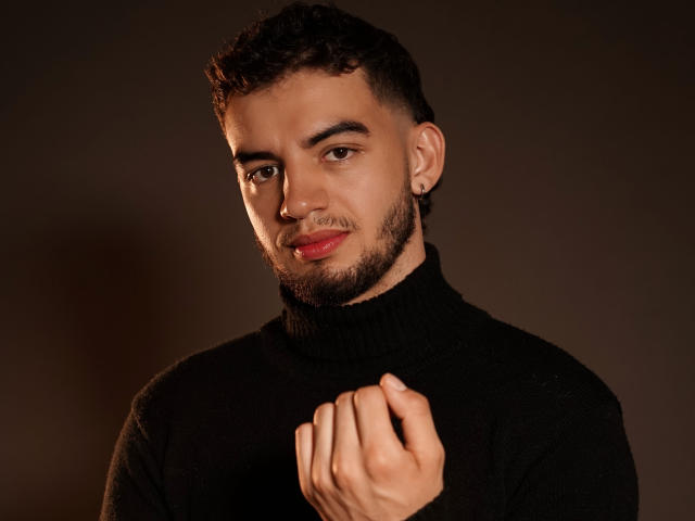 MichaelMagno with curly dark hair, beard, and nose ring, wearing a black turtleneck, holding his chest with a slight smile, brown eyes, and dark skin.