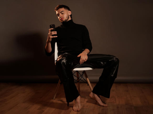 MichaelMagno is barefoot, wearing a black turtleneck and shiny black pants, sitting on a white chair, holding a phone, with a beard and short curly hair, looking down.