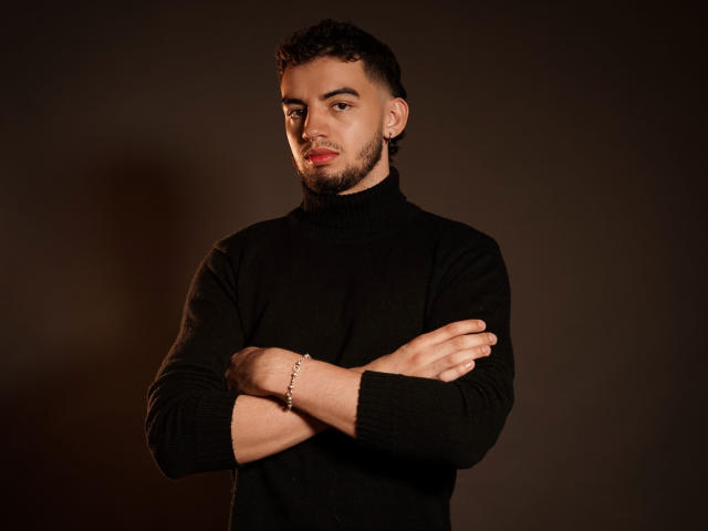 MichaelMagno, with a dark beard and short curly hair, stands with arms crossed, wearing a black turtleneck, looking slightly annoyed, and has a silver bracelet on his right wrist.