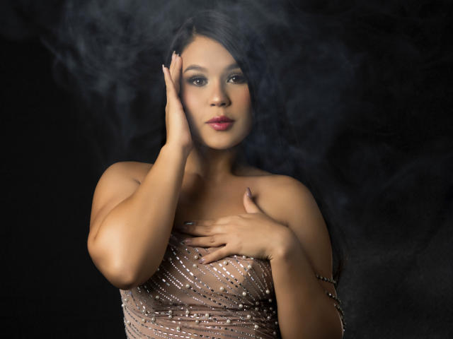CarolineWill with smoky background, hand on cheek, other hand on chest, wearing sheer, sparkly dress, brown skin, dark hair, pink lipstick, looking slightly to the side, seductive.
