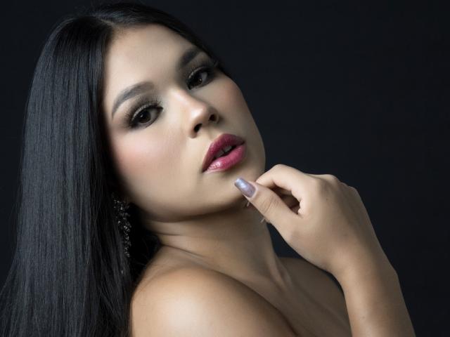 CarolineWill with long black hair, dark eyes, pink glossy lips, touching her bottom lip with a manicured finger, wearing dangling earrings, against a dark background.