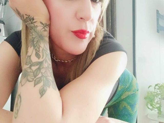 SammyDesire, con le labbra rosse, la camicia nera e il vestito a motivi verdi, appoggia il braccio tatuato su un tavolo, con la testa appoggiata alla mano, indossando una collana a catena, in una stanza luminosa..