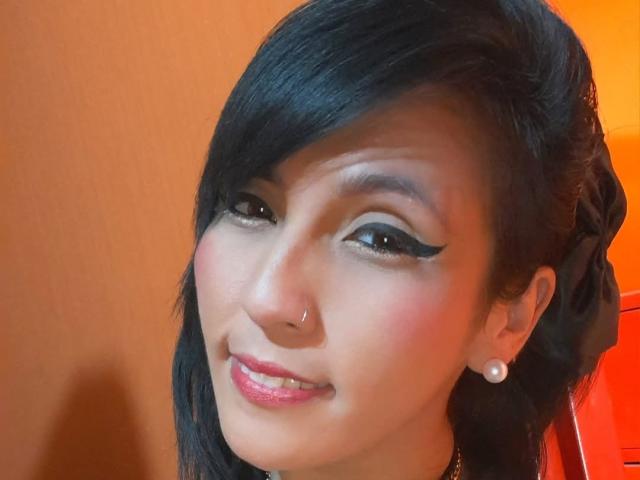 DaniaHot69 hat schwarzes Haar, einen Nasenring und Perlenohrringe, trägt auffälligen schwarzen Eyeliner, pinken Lippenstift und hat ein leichtes Lächeln, wodurch sie süß und sexy aussieht..