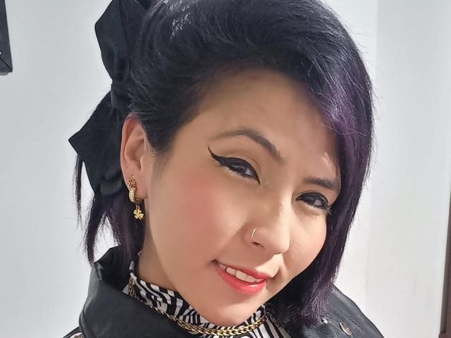 DaniaHot69 a les cheveux d'un violet foncé, un nœud noir, et porte des boucles d'oreilles en forme d'étoiles dorées. Elle sourit, avec du rouge à lèvres, du crayon noir pour les yeux, et un anneau de nez. Elle porte un haut à rayures zébrées..