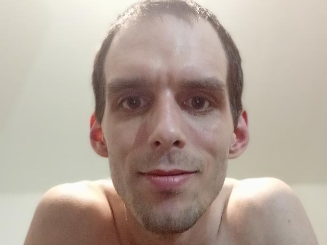 Shirtless Qeal69 with short brown hair, light skin, slight stubble, and a subtle smile, looking directly at the camera with slightly wet skin.