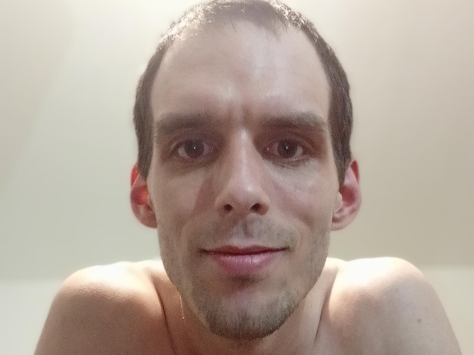 Shirtless Qeal69 with short brown hair, light skin, slight stubble, and a subtle smile, looking directly at the camera with slightly wet skin.