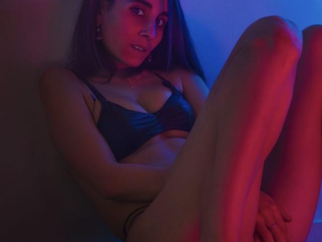 WildShowBeauty with dark hair, wearing a black bra, sitting with legs up, illuminated by blue and red lights, showing her tits and a slight smile, hands on her knees.