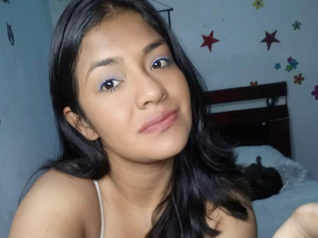 ArianithaXox, avec ses cheveux noirs, son eye-liner bleu et ses lèvres roses, portant un haut blanc, assise sur un lit, regardant légèrement sur le côté avec un sourire discret..