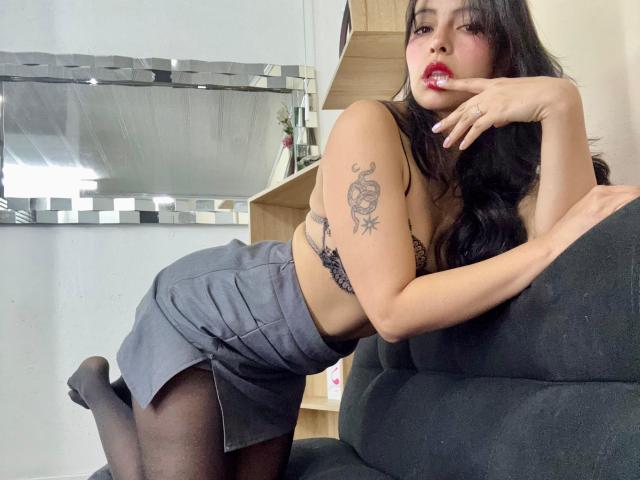 EliieRuizz with long black hair, red lipstick, and tattoos, leaning on a dark chair, wearing a gray skirt, black bra, and sheer stockings, biting her finger.