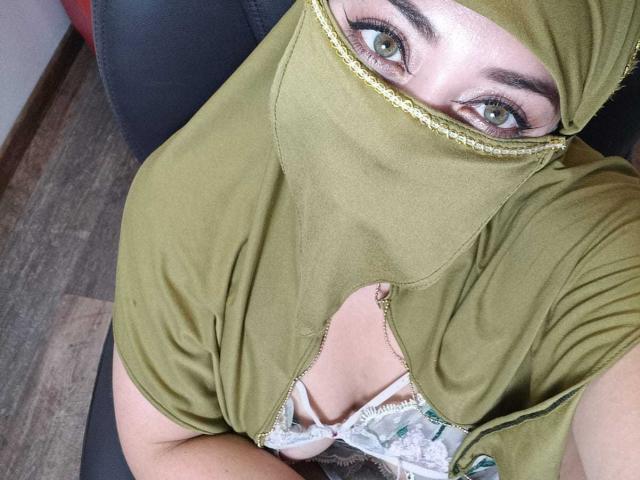 ZahraJalid with olive-green mask covering her face, green eyes looking up, olive-green top with cutout exposing white and green lace bra, sitting on dark chair, wood floor.