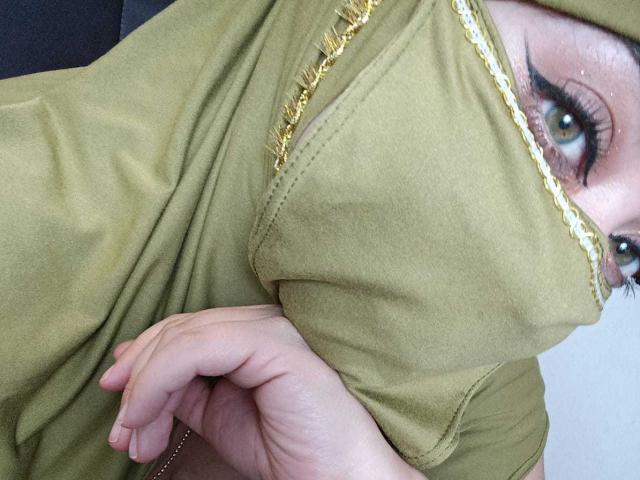 ZahraJalid's face with green shirt covering her tits, showing one eye with thick black eyeliner, her hand near her chest, and a gold-trimmed edge of the shirt.