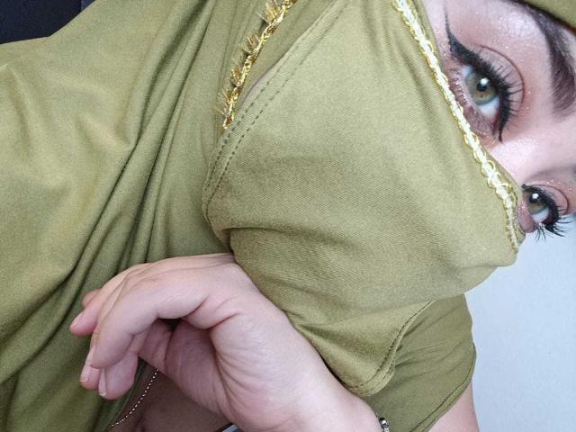 ZahraJalid's face, olive-green hijab, and hand in frame, showing green eyes with thick eyelashes, and her hand clutching the fabric. Her gold necklace is visible.
