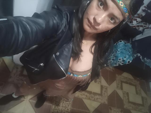 ArianithaXox con un vestito marrone, una giacca di pelle nera, una fascia colorata e i seni visibili, che si fa un selfie, guardando verso l'alto, in una stanza con piastrelle, indossando stivali neri..