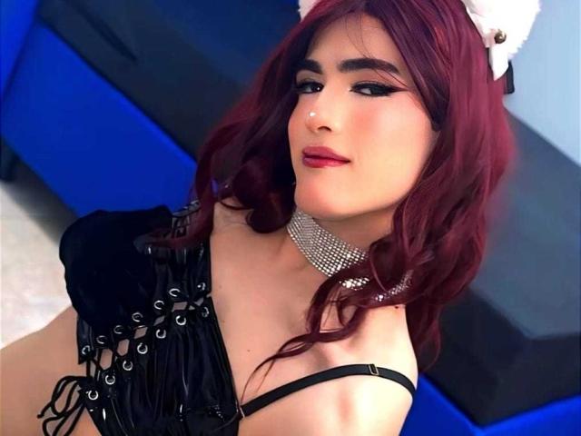 MichellPratTrans has red hair, cat ears, black lace-up corset, and a sparkly choker. She's looking up with eyeliner and red lipstick. Boobs are partially visible.