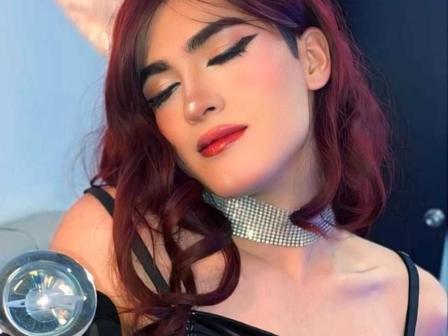 MichellPratTrans with red lipstick, closed eyes, black eyeliner, shiny skin, and a diamond choker, has dark red wavy hair, black top, and a cute, confident expression. Boobs visible.