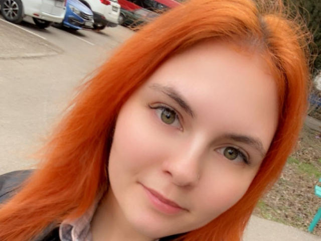 LexhamMoom has bright orange hair, green eyes, and a light skin tone, smiling slightly with a cute face and subtle makeup, wearing a gray shirt in a selfie.