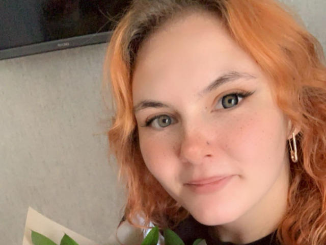 LexhamMoom has light green eyes, wavy orange hair, and is smiling softly with a slight freckle on her nose. She's wearing small gold earrings and has a light skin tone.