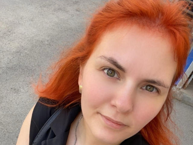 LexhamMoom has bright red hair, green eyes, and a slight smile. She's wearing a black shirt and gold earrings. Her ass is not visible. She's looking at the camera.