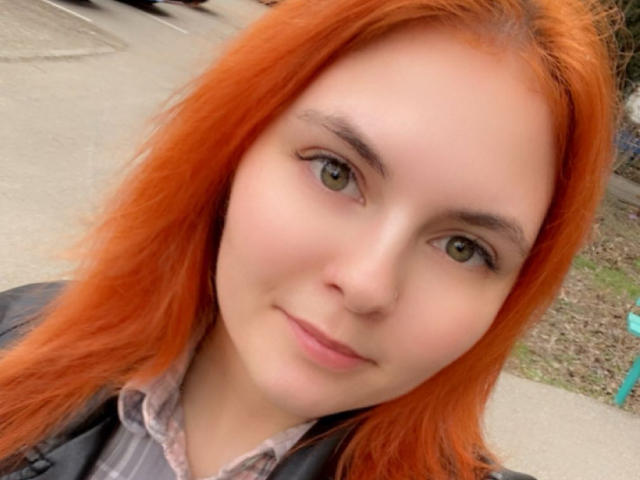 LexhamMoom has bright orange hair, green eyes, and a cute face with a slight smile. She's wearing a gray shirt and black jacket. Her tits are not visible.