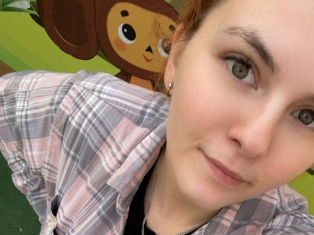 LexhamMoom has pale skin, green eyes, and wears a pink and gray plaid shirt, with a slight smile, and small earrings, in a selfie with a cute monkey face.
