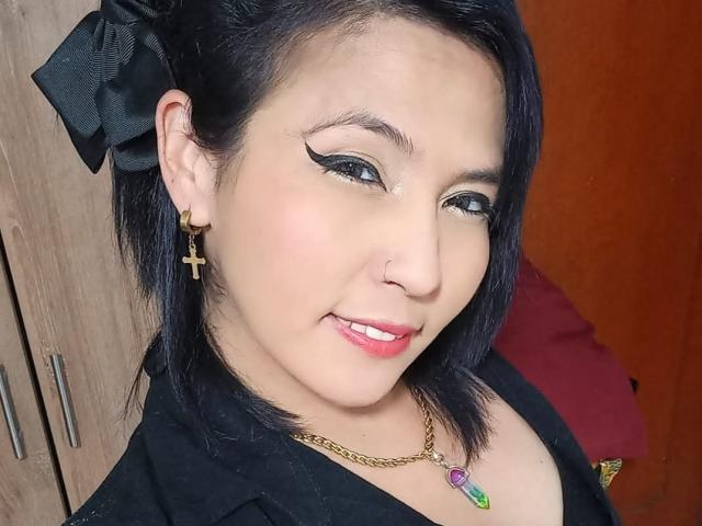 DaniaHot69 has black hair with a bow, black eyeliner, a nose ring, cross earrings, and a gold necklace with a green and purple gem. She's smiling with pink lipstick.