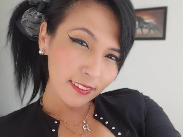 DaniaHot69 ha i capelli neri raccolti in una coda di cavallo, indossa una maglietta nera con borchie argentate, orecchini di diamanti e una collana d'oro con un ciondolo a forma di cuore. Sorride, con il rossetto rosso..