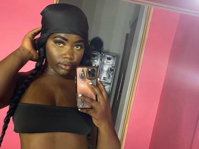 SweeterChocolate with dark skin, braided black hair, black headscarf, black strapless top, holding pink phone, pink wall, mirror selfie, looking at camera, slightly parted lips.