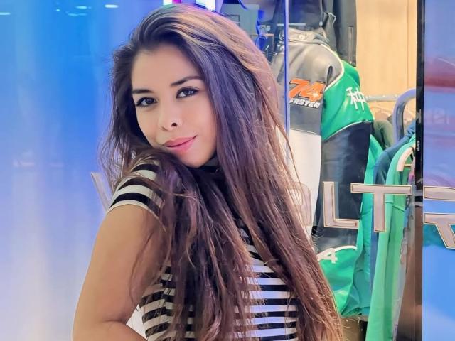 AyeBeauty with long brown hair, striped shirt, and a slight smile, looking over her shoulder, sitting in a room with sports gear and a green chair.