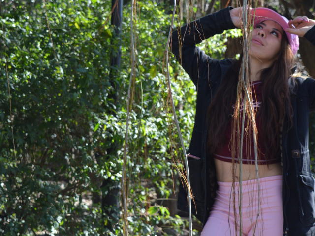 AyeBeauty with long brown hair, pink cap, black jacket, red crop top, and pink pants, standing in sunlight, hand on cap, looking up, surrounded by hanging vines.