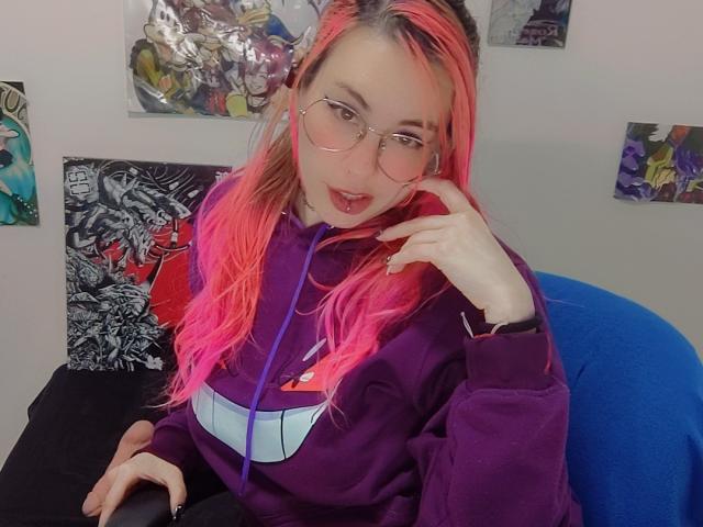 NatashaLau with pink and orange hair, wearing a purple hoodie, glasses, and a tongue-out expression, touching her cheek. She's sitting on a blue chair, looking cute.