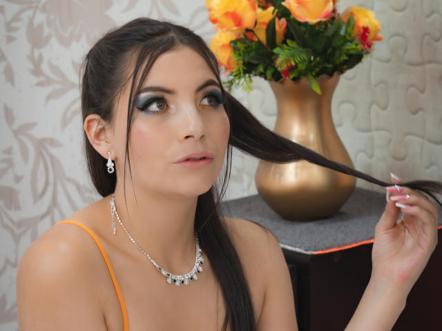 ValleryCortez with dark hair, holding a hair strand, wearing orange top, silver necklace, and earrings, looking up, orange and yellow flowers in a gold vase in the background.