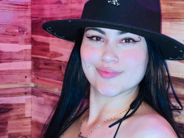 EmiliaCartersh met zwarte hoed, neusring en eyeliner, lachend met roze lippen, waarbij kleine borsten zichtbaar zijn, met lang zwart haar en een ketting, op een houten achtergrond..