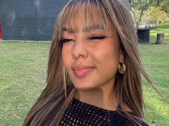 AmelieDuvall with brown, wavy hair and bangs, nose ring, and gold earrings, smiling with pink lipstick, wearing a black mesh top, outdoors on a grassy area.