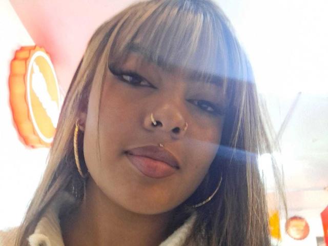 AmelieDuvall has a nose ring and gold hoop earrings, sticking out her tongue with long, straight brown hair and heavy eyeliner. Sun flare in the background.