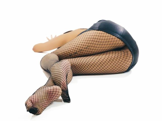 AdelaRanger lying on her side, wearing black fishnet stockings and a tight black leather mini-skirt, with her ass and feet prominently in the foreground.